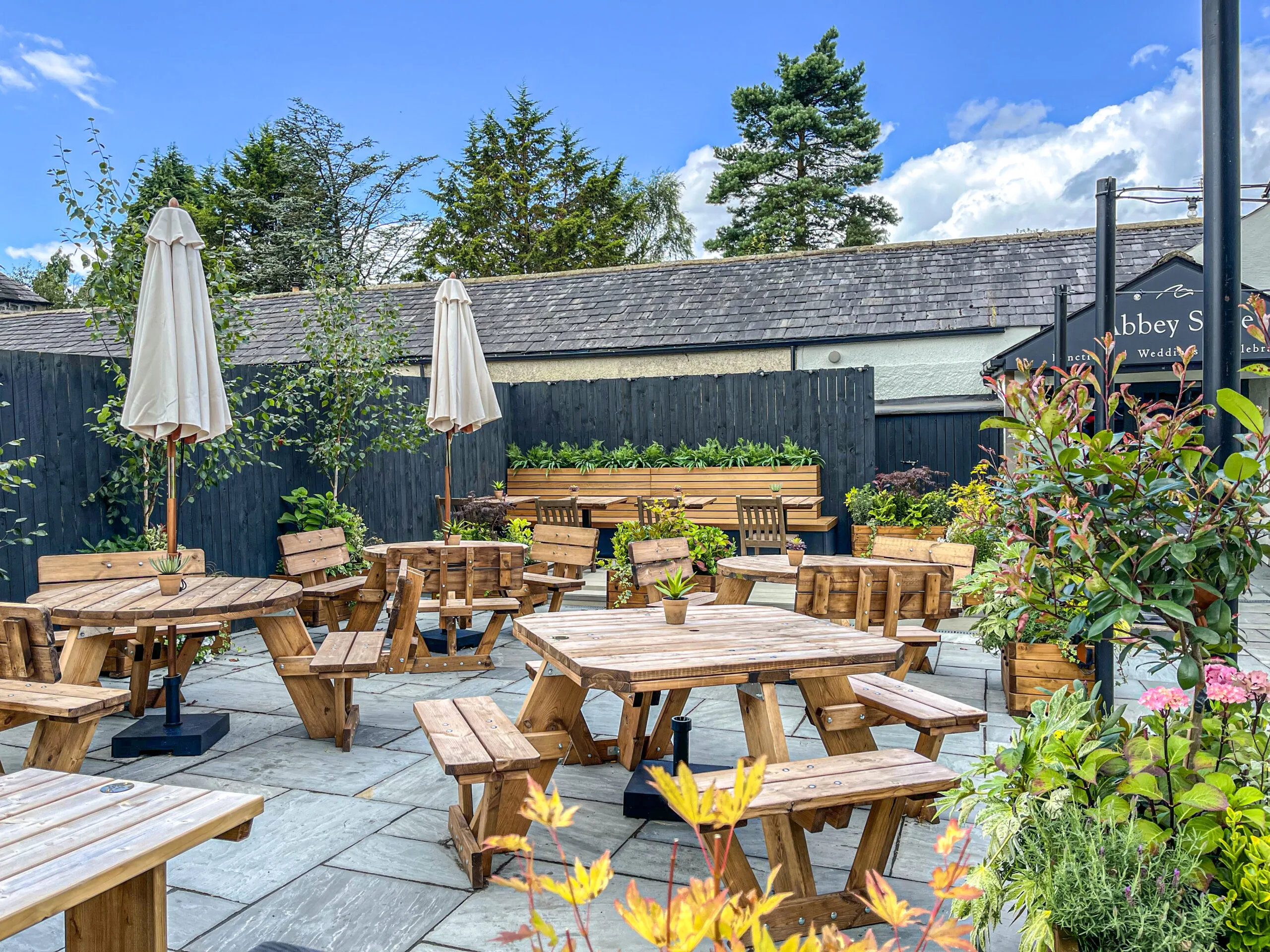 A beautiful courtyard at the Spread Eagle Inn in Sawley, surrounded by greenery and mature trees, with rustic wooden picnic tables, benches and cream parasols. The courtyard provides an outdoor space for wedding and event guests, while also serving as a relaxed beer garden when events are not taking place. A traditional slate-roofed building and Abbey Suite wedding signage can be seen in the background, creating a charming countryside setting.