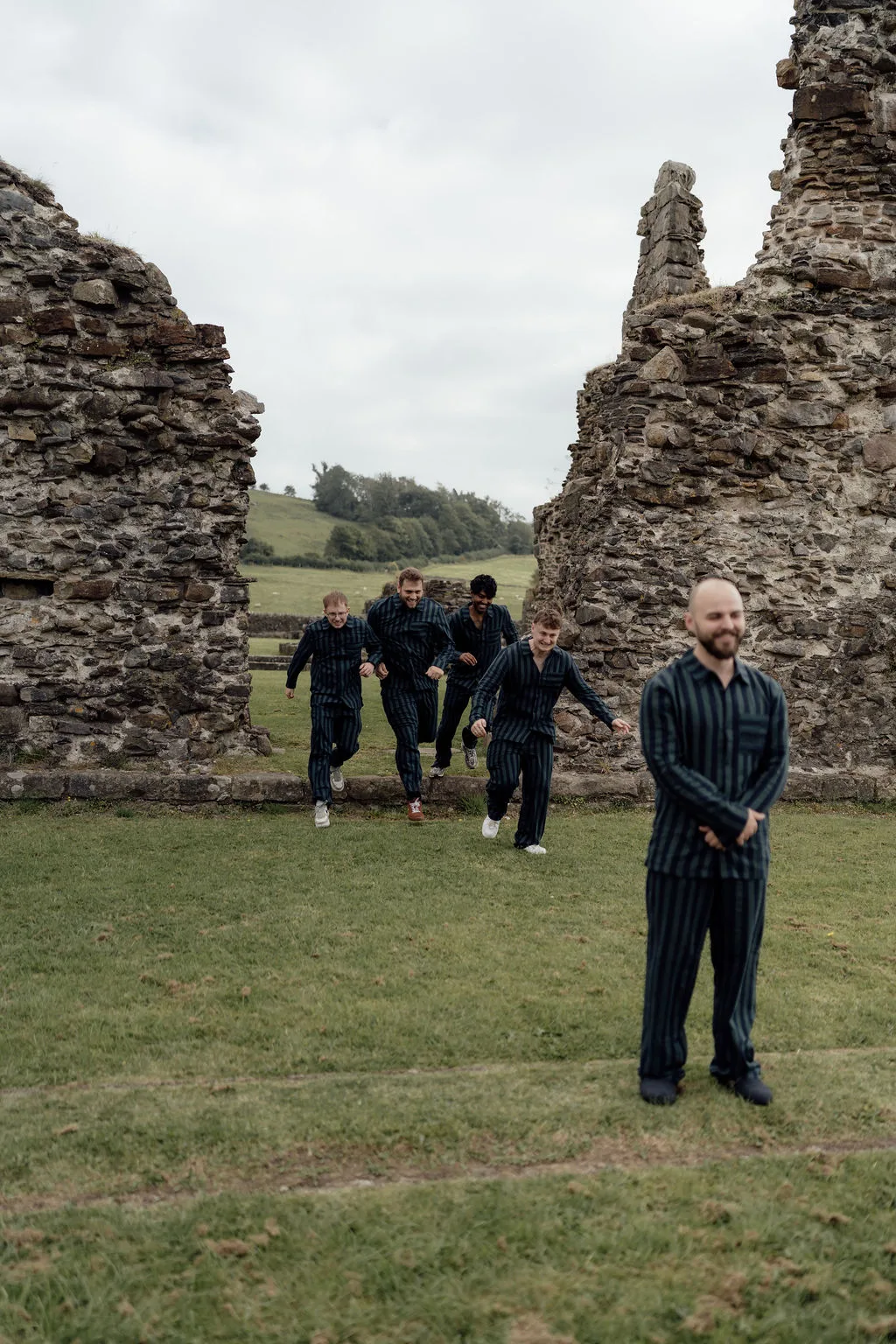 A group of groomsmen in matching dark striped suits walking through the historic ruins of Sawley Abbey, surrounded by open green countryside and rolling hills. The ancient stone ruins provide a dramatic backdrop, capturing a relaxed and playful moment during a wedding at the Abbey Suite at the Spread Eagle Inn in Sawley.