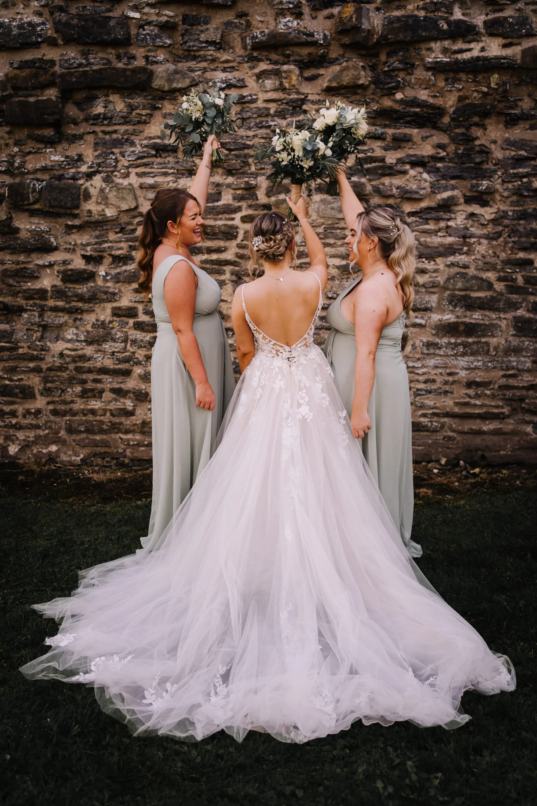 A bride stands with her back to the camera between two laughing bridesmaids in sage green gowns, all three raising bouquets of white roses and eucalyptus in the air. The bride's dress has an open lace back and a long tulle train spread across the grass, set against an old weathered stone wall.