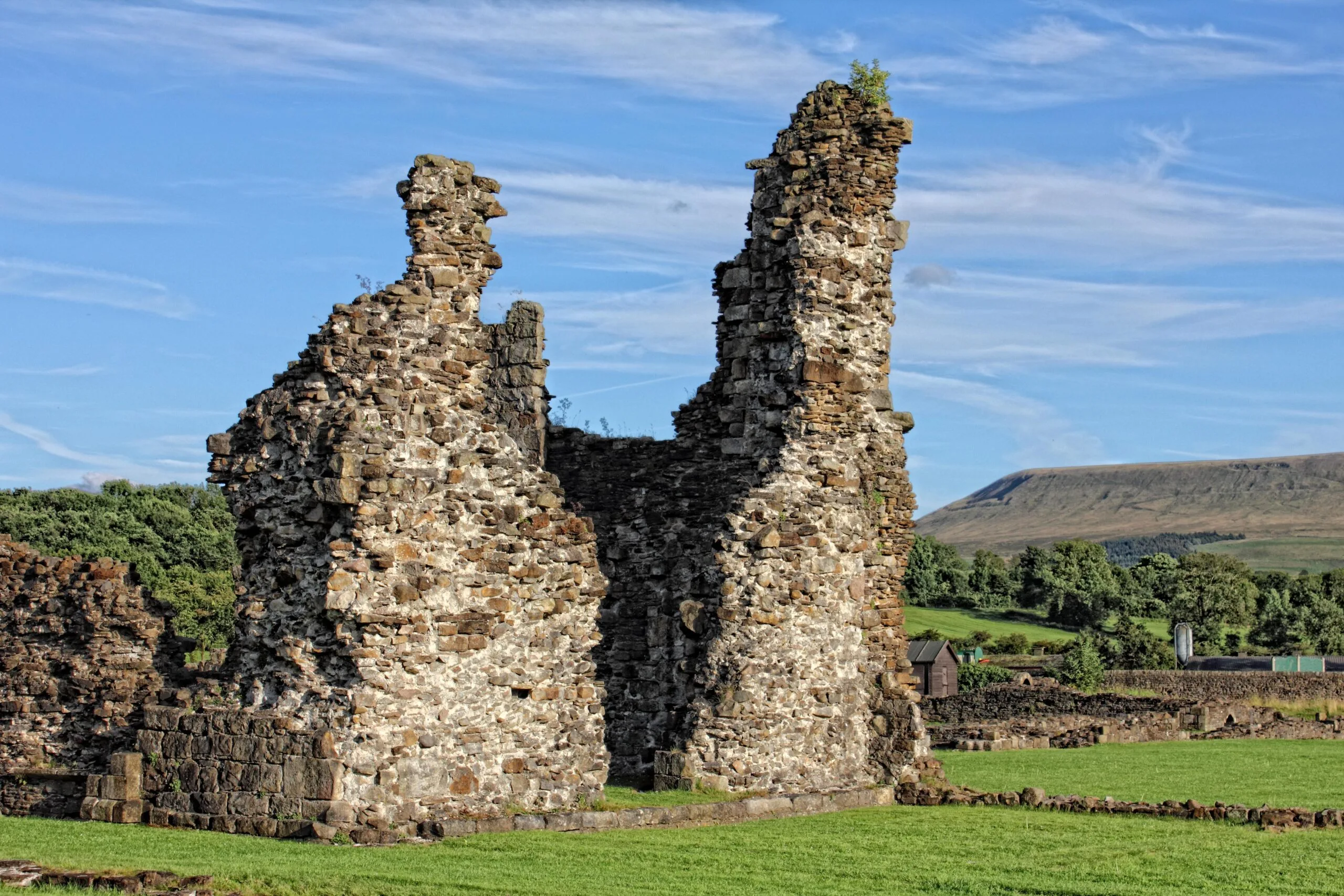 The ruined stone walls of Sawley Abbey standing tall on a sunny day, with two jagged towers of weathered stonework rising against a blue sky streaked with thin cloud. Green lawns and low foundation walls surround the ruins, with trees and Pendle Hill in the distance.